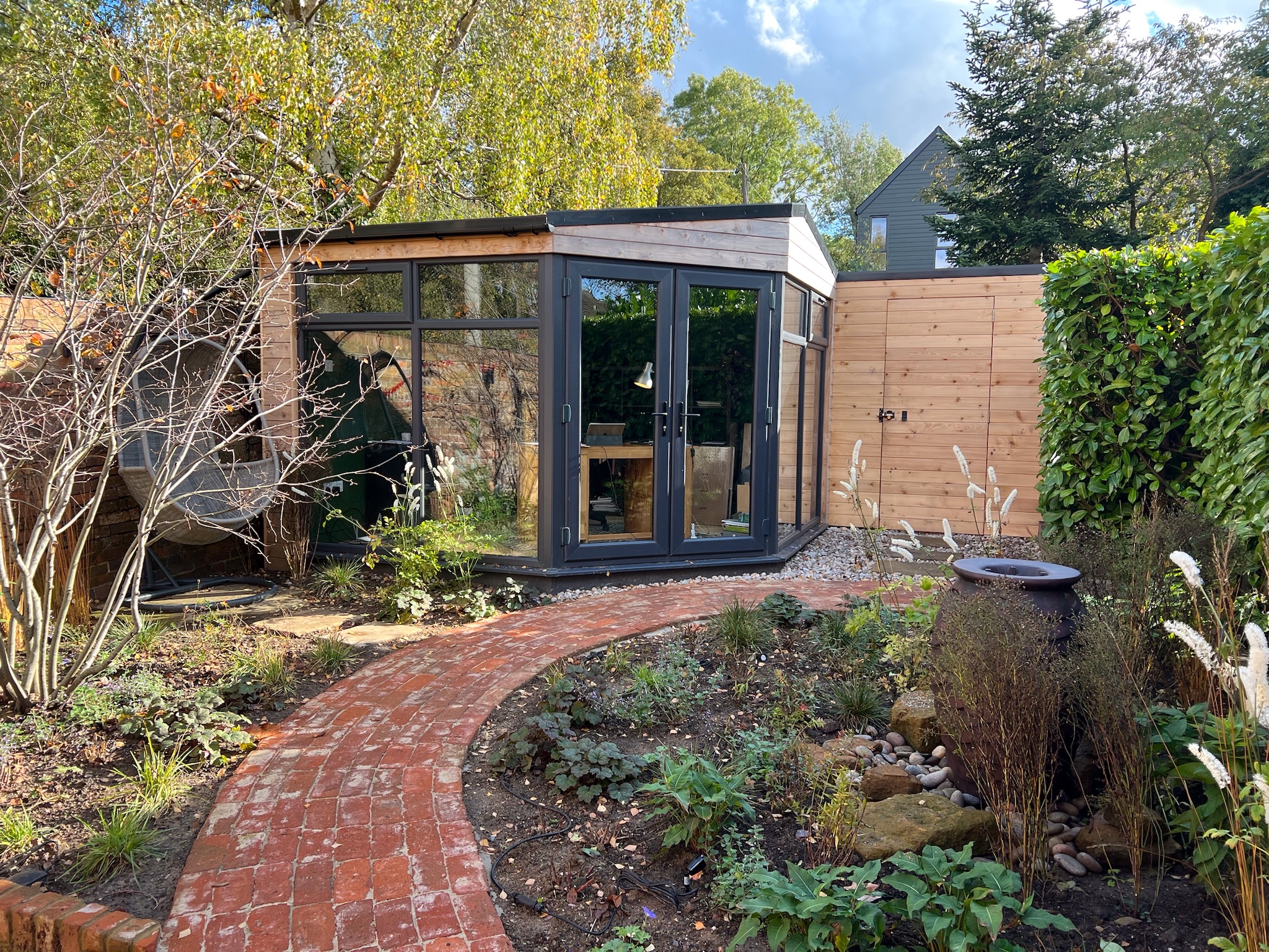 Reclaim brick path surrounded by autumnal planting leading to a timber clad out door office